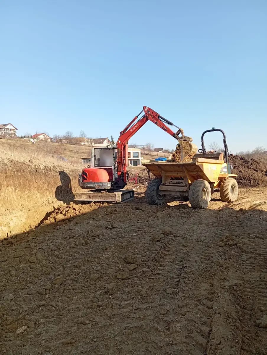 Excavator care încarcă pământ direct într-un dumper, pe un teren săpat la marginea localității