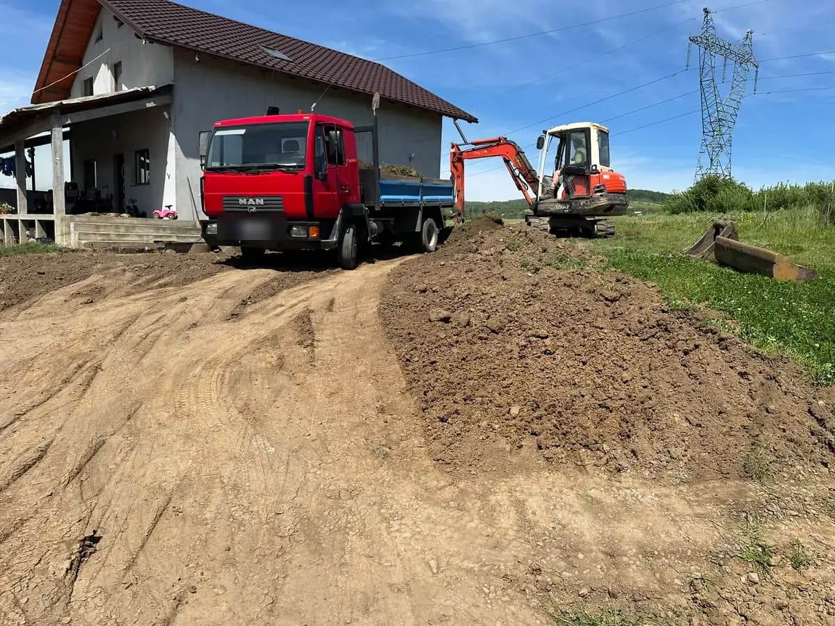 Camion basculant încărcat cu pământ și excavator pe șenile, la nivelarea unui teren de lângă o casă