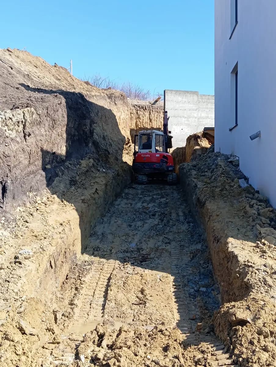 Șanț săpat cu excavatorul între malul de pământ și peretele unei clădiri, pe un culoar îngust