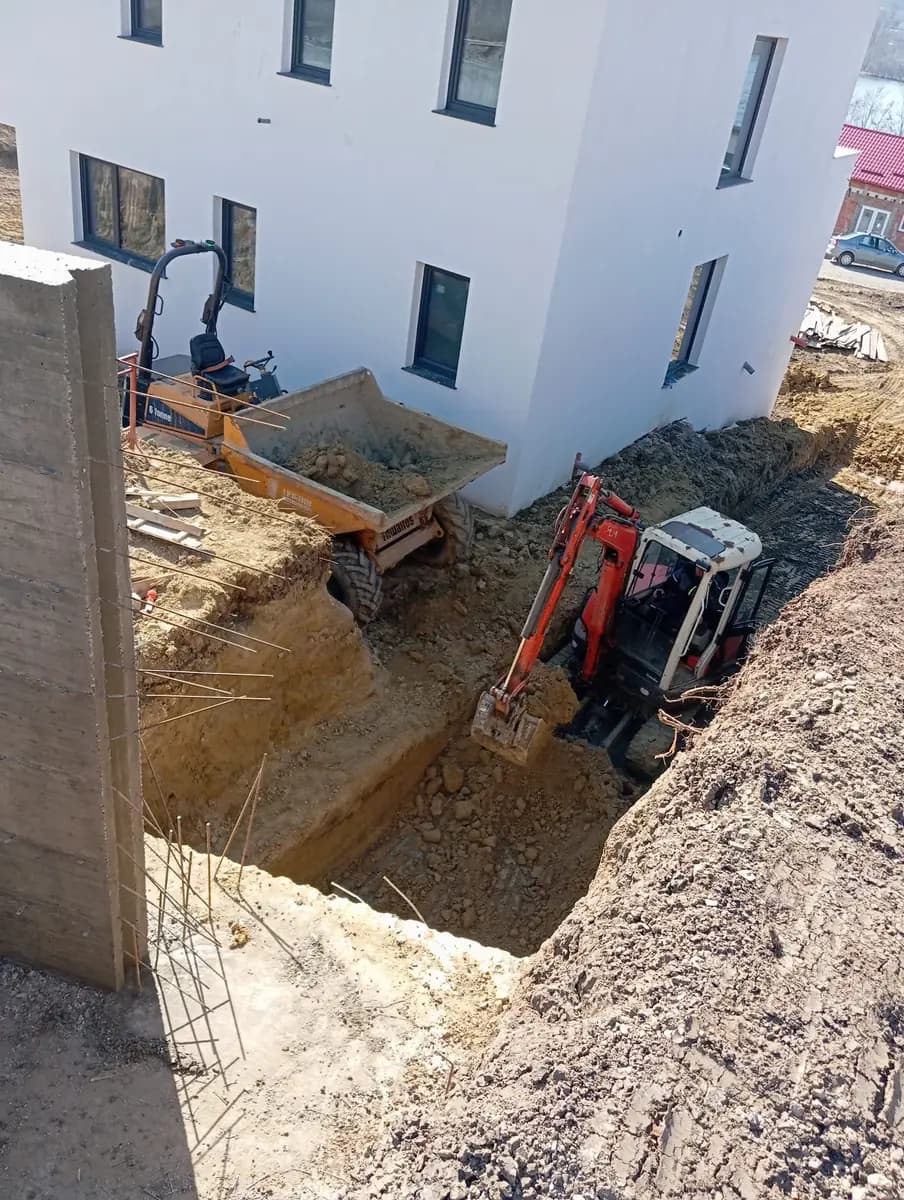 Săpătură făcută cu excavatorul lângă peretele unei clădiri noi, cu pământul încărcat direct în basculantă