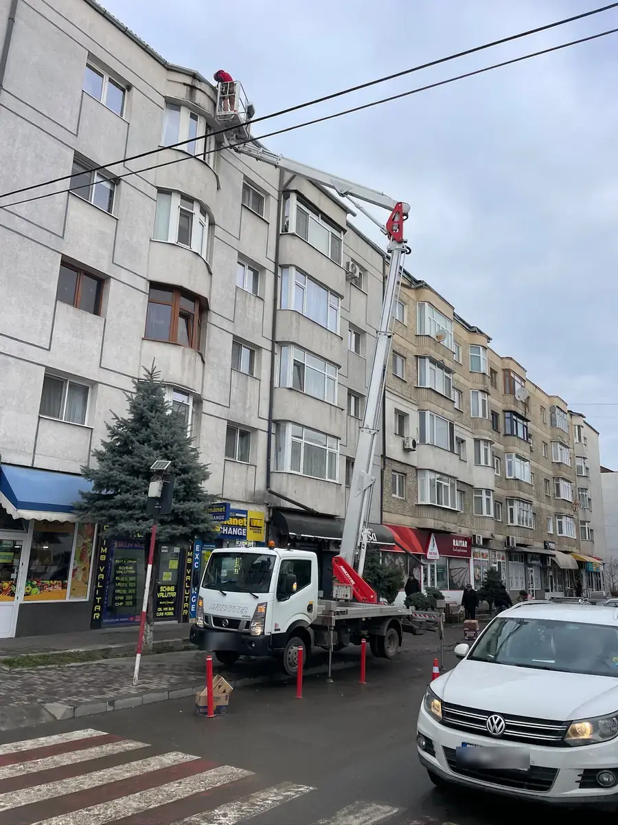 Nacelă cu brațul ridicat la ultimele etaje ale unui bloc cu spații comerciale la parter, pe o stradă cu trafic